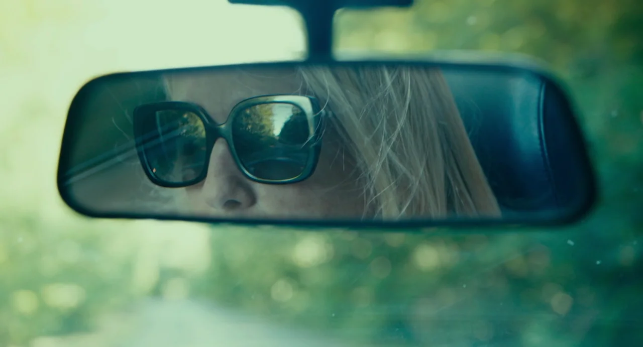 Last Summer (2023) movie still: closeup — The rearview mirror reflects a woman with blonde hair wearing dark sunglasses while driving. Through the…