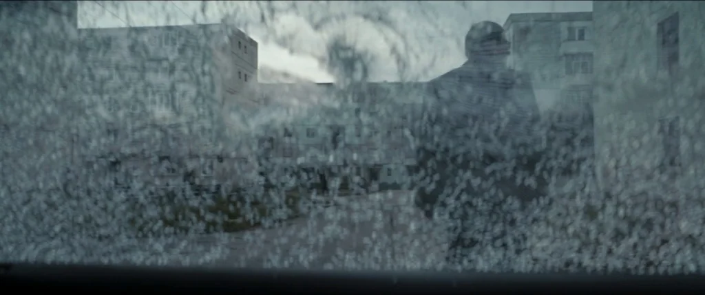 Graduation (2016) movie still: wide — A view through a shattered glass pane onto a cluster of grey apartment buildings. The foreground is heavily…