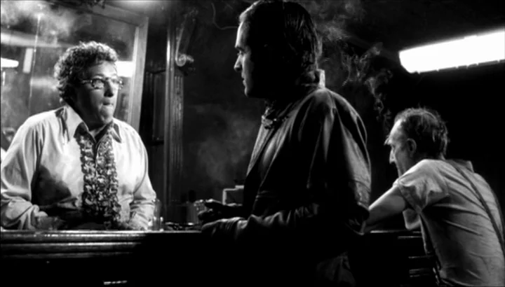 The American Astronaut (2001) movie still: medium — A dark, smoky bar interior with a reflective surface behind the counter. Two men talk while…