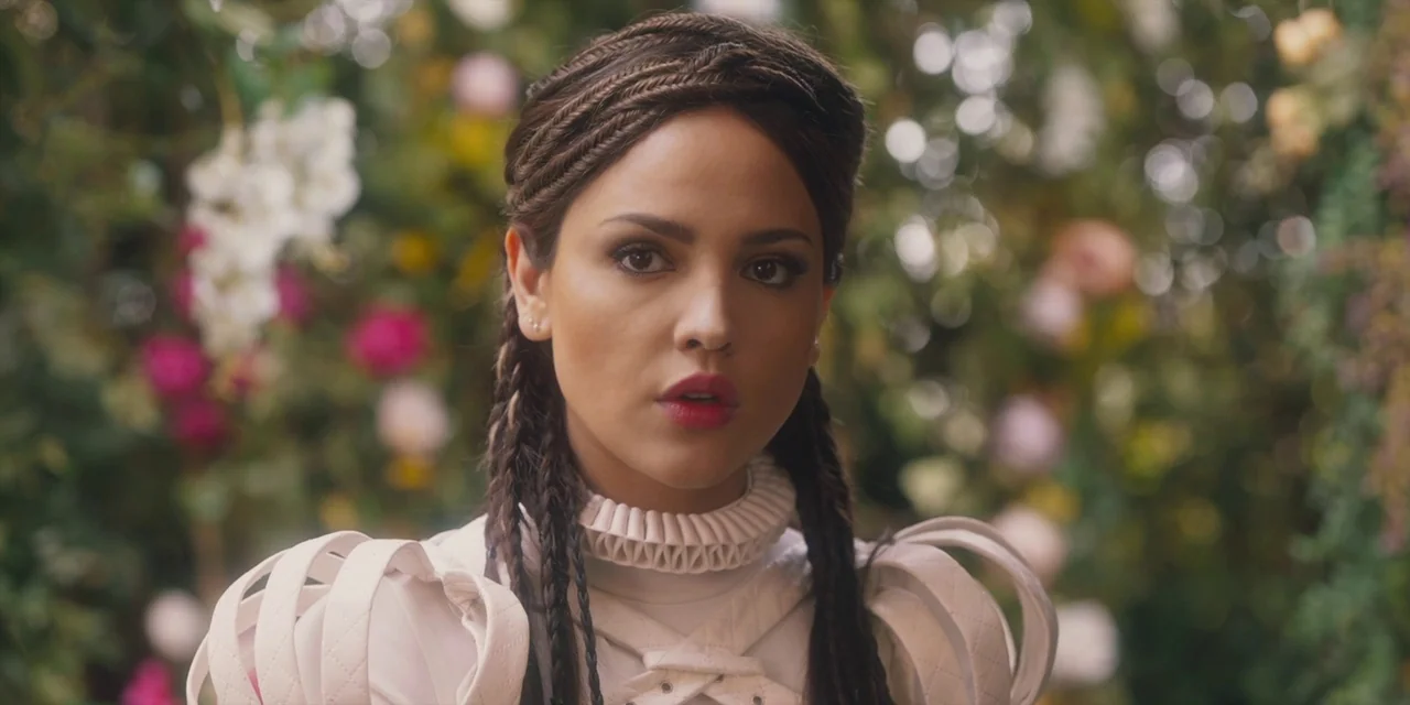 Paradise Hills (2019) movie still: closeup — A woman stands in front of a dense floral display filled with various blossoms. She is centered in the…