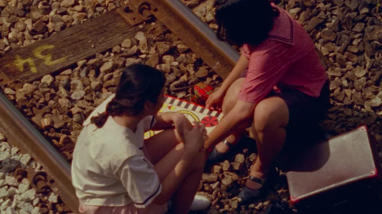 Shirkers (2018) movie still: medium — A pair of train tracks with wooden ties rests on a bed of loose gravel. Two young women sit between the tracks…