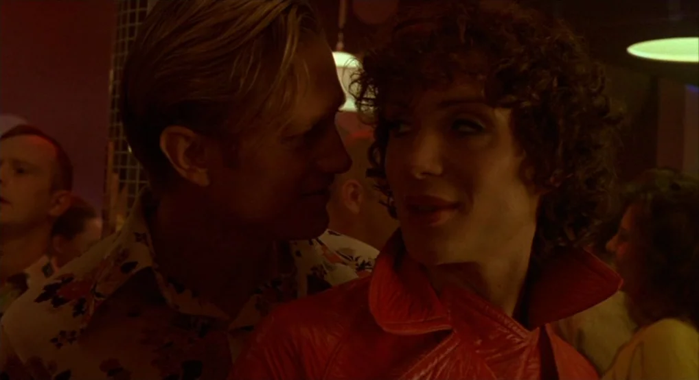 Breakfast On Pluto (2005) movie still: medium — A crowded nightclub setting with low light and red neon accents. A blonde man whispers something…