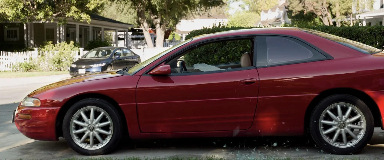 Ted 2 (2015) movie still: medium — A red coupe sits parked on a residential street with its driver-side window smashed. Shards of glass scatter onto…