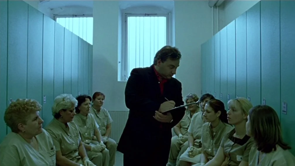 Import Export (2007) movie still: medium — A narrow room is lined with tall lockers on two sides and a central window. A man in a dark suit stands…