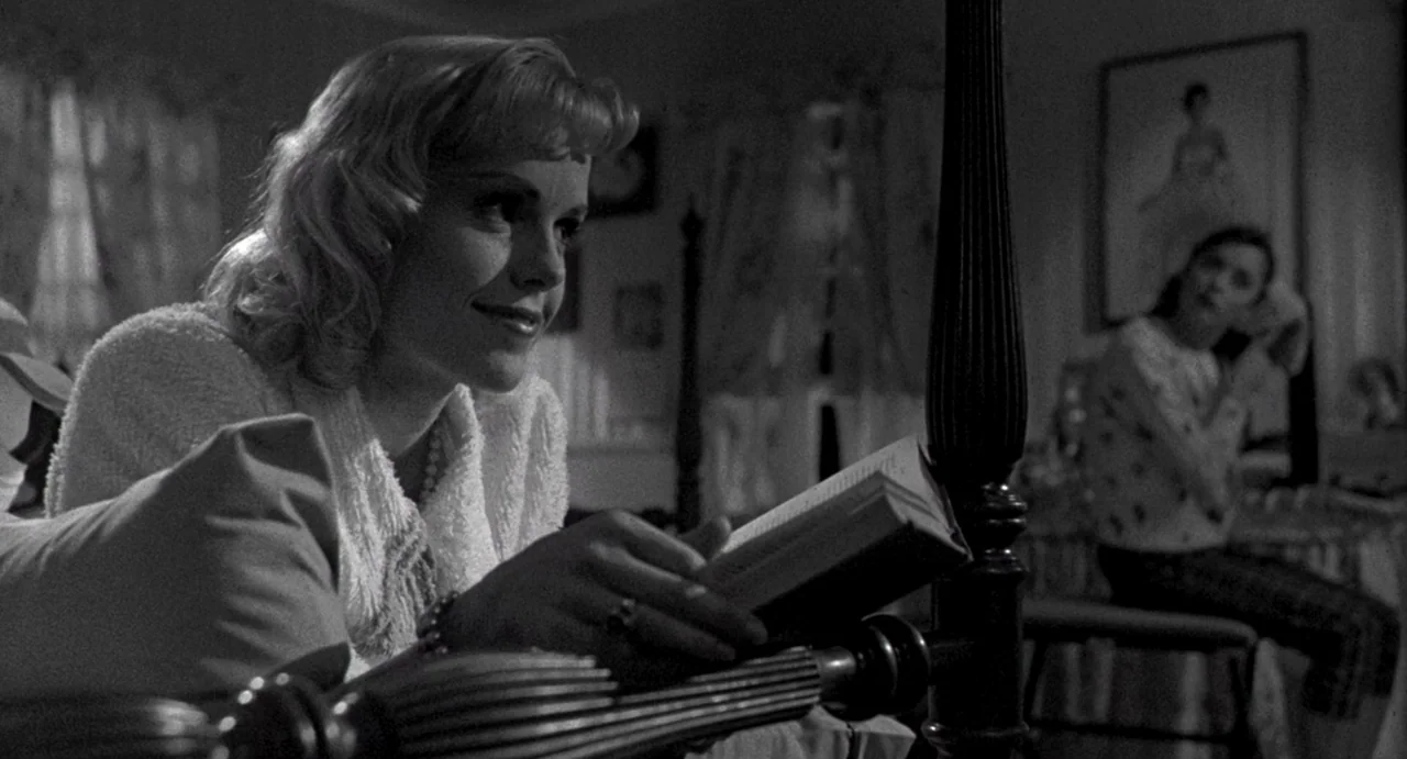 Night of the Creeps (1986) movie still: medium — A dimly lit room with a wooden bed frame and a wall-mounted picture frame. One woman reads a book…