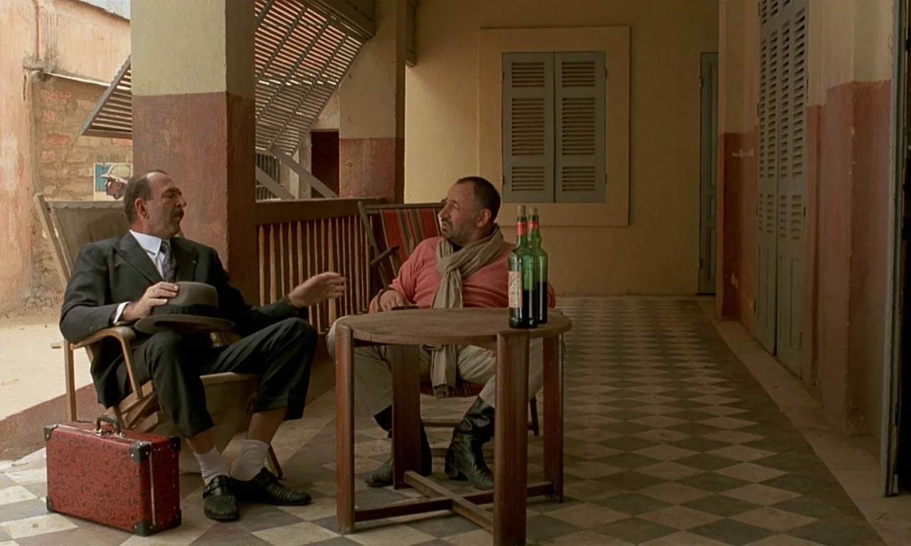 Coup De Torchon (1981) movie still: fullbody — A shaded colonial-style porch has checkered tile floors and faded rust-colored wall trim. Two…