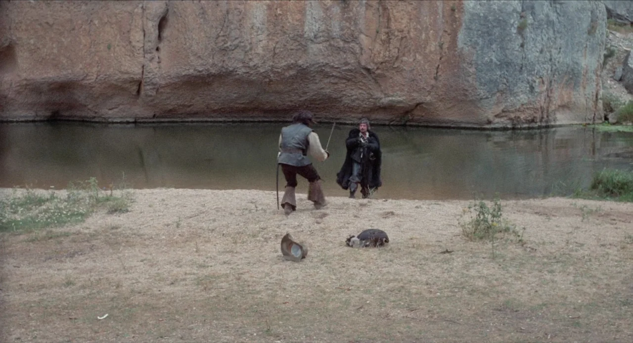 The Three Musketeers (1973) movie still: wide — A sandy riverbank sits before a sheer, eroded sandstone cliff face at the water's edge. Two men in…