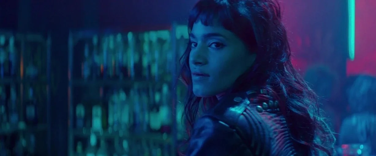 Atomic Blonde (2017) movie still: closeup — A dimly lit bar interior features rows of blurred bottles against blue-lit shelves. A young woman in a…
