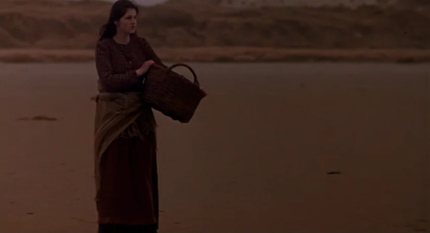 The Secret Of Roan Inish (1994) movie still: fullbody — A woman stands on a barren, sandy shoreline during the late evening light. She holds a woven…