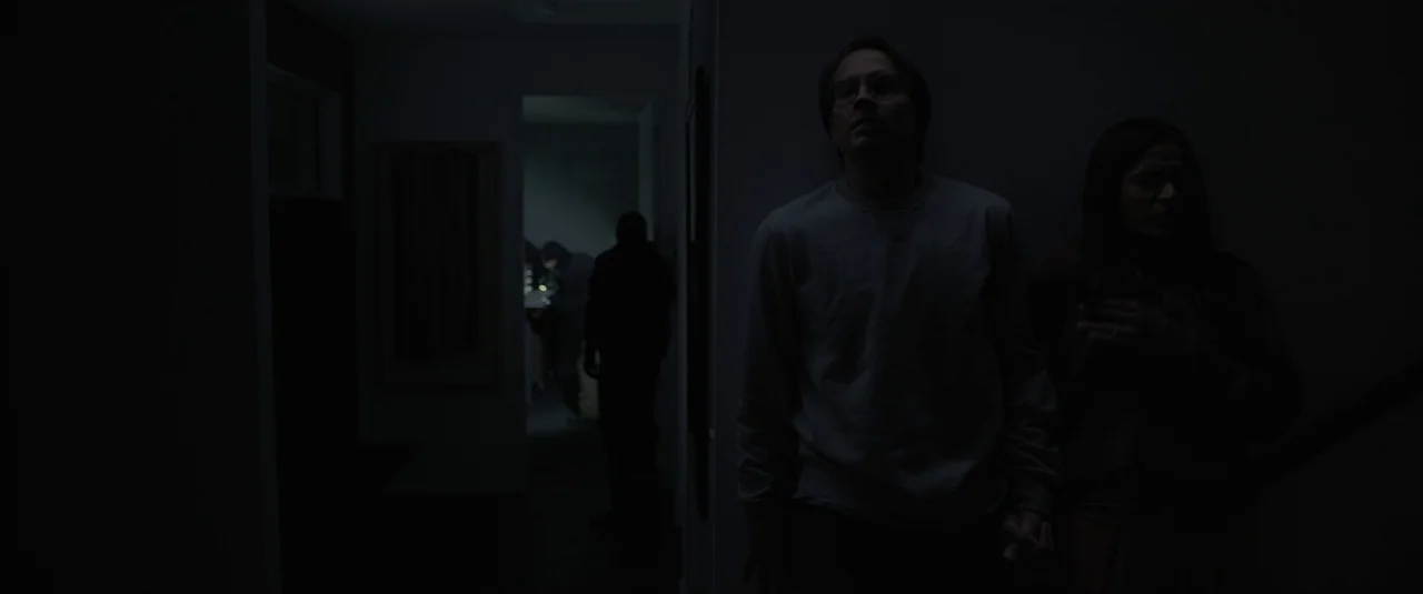 Intrusion (2021) movie still: medium — A dark residential hallway with a man and woman peering toward a distant opening. A silhouette of a third…