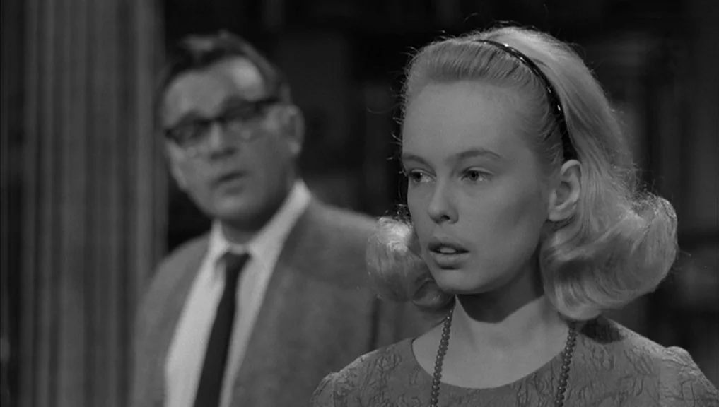 Who’s Afraid Of Virginia Woolf? (1966) movie still: medium — A dark indoor space where a woman stands in the foreground looking away. A man in…