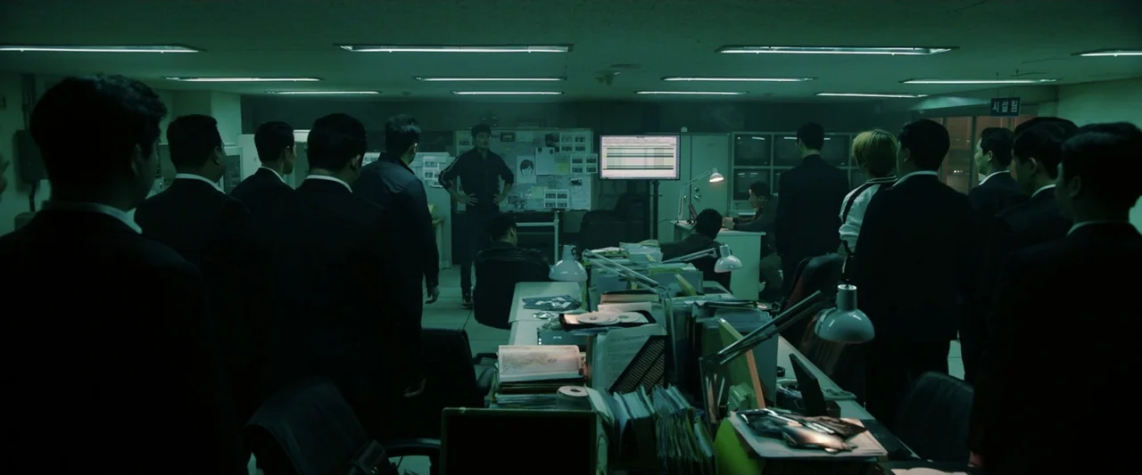 The Gangster, the Cop, the Devil (2019) movie still: wide — A dark office space is filled with multiple men in professional dark business suits. The…