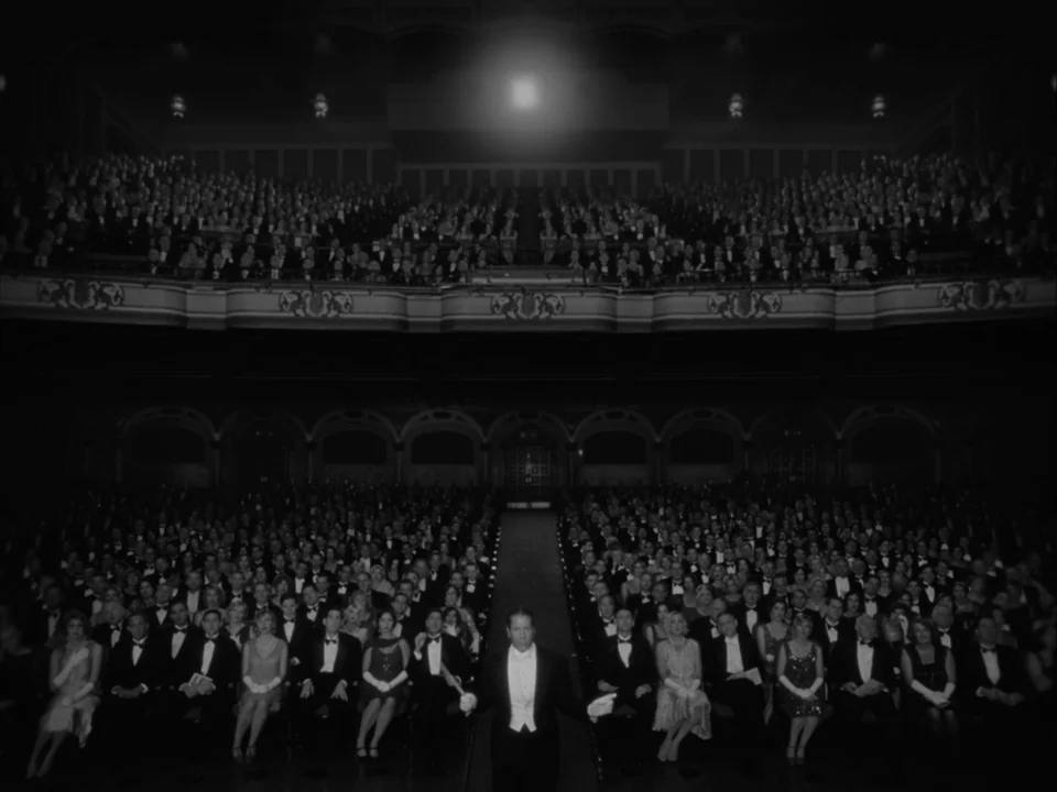 The Artist (2011) movie still: wide — A crowded 1920s-style theater auditorium with an audience seated in formal evening wear. A conductor stands…