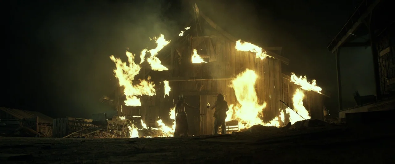 The Lone Ranger (2013) movie still: wide — A wooden building is engulfed in flames at night in a desolate rural area. Two figures stand facing the…