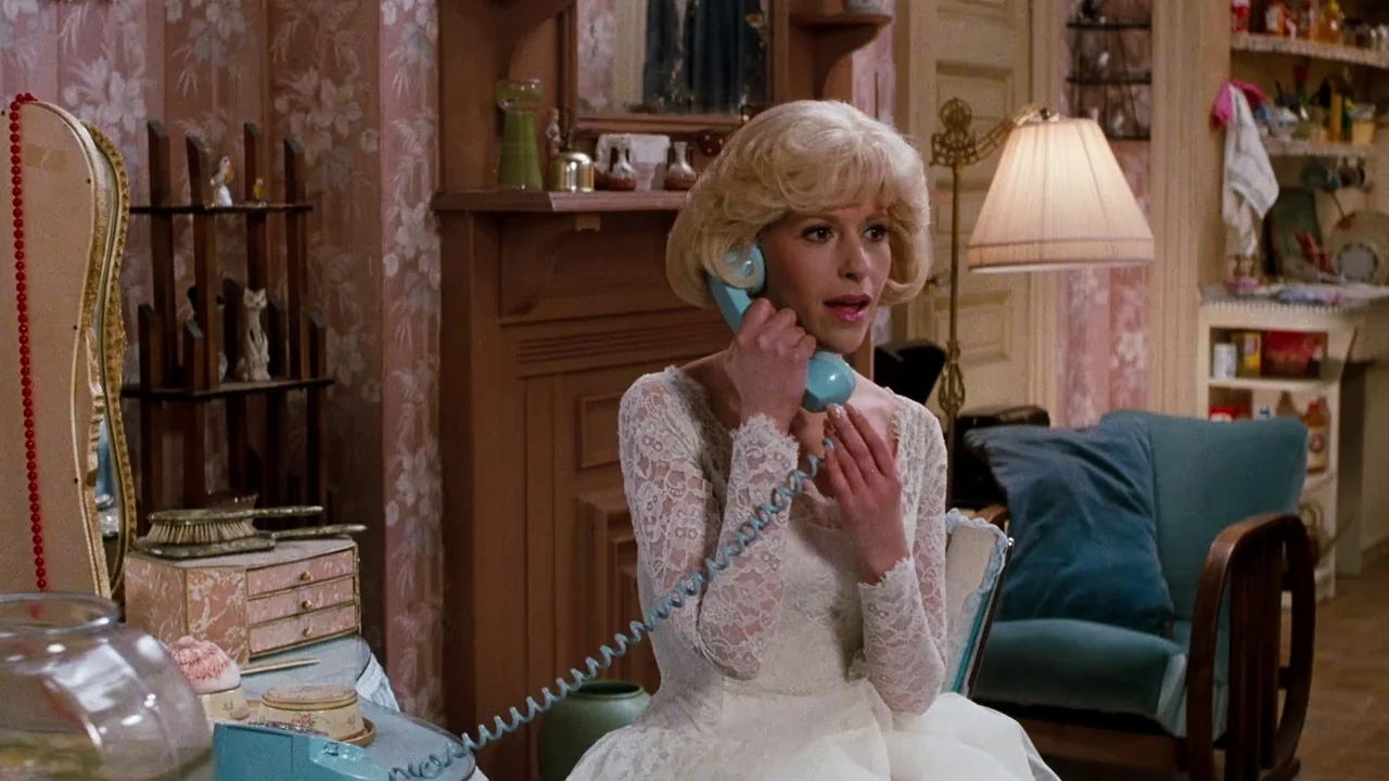 Little Shop of Horrors (1986) movie still: medium — A vintage-inspired room with floral wallpaper and a wooden fireplace mantle. A woman in a white…