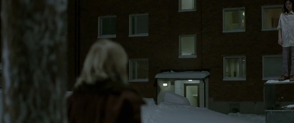 Let The Right One In (2008) movie still: medium — A dark brick apartment building stands behind a snow-covered yard at night. A girl stands on an…