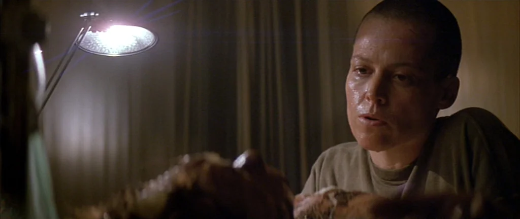 Alien 3 (1992) movie still: medium — A dimly lit room with simple, industrial-style curtains visible in the background. A woman with a shaved head…