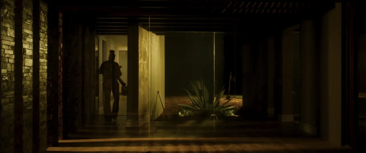 Phobias (2021) movie still: fullbody — A dark, modern house interior features a large glass wall looking out into a courtyard. A man stands in the…