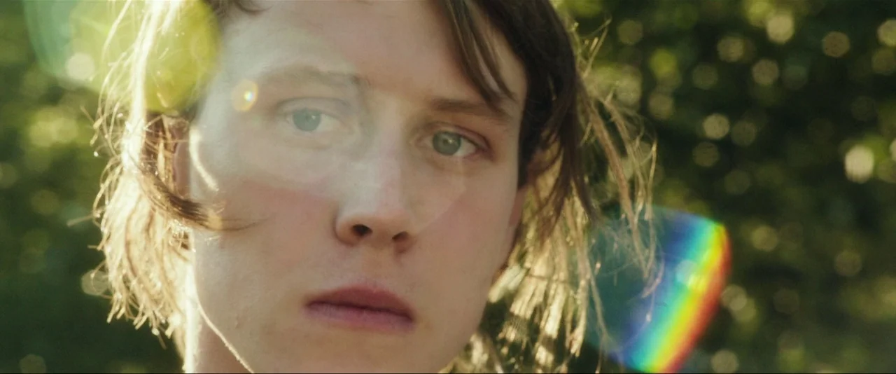 Captain Fantastic (2016) movie still: closeup — A person is positioned against a background of out-of-focus green foliage. Intense golden sunlight…
