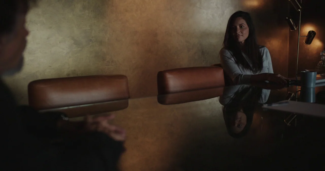 Violet (2021) (2021) movie still: medium — A dimly lit room features a textured bronze wall and a large reflective glass table. A woman sits on the…
