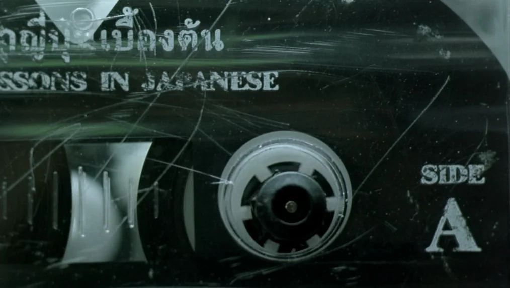 Last Life in the Universe (2003) movie still: closeup — A close-up view of a black cassette tape with heavy surface scratches. The text 'LESSONS IN…
