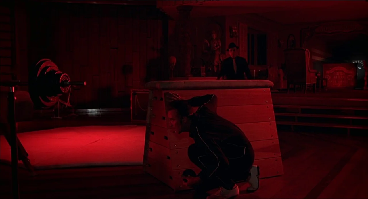 The Man With the Golden Gun (1974) movie still: wide — A dimly lit, red-saturated gymnasium interior contains a lone wooden vaulting horse. A man…