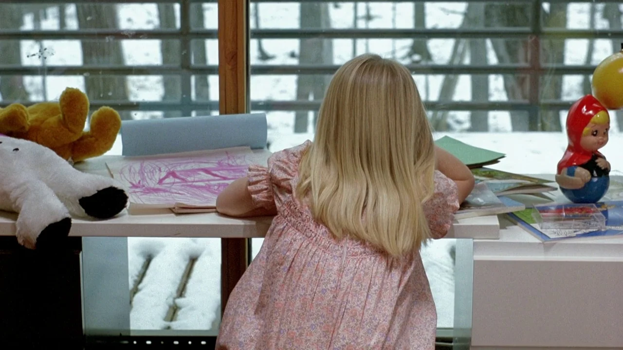 The Brood (1979) movie still: medium — A white desk sits in front of a floor-to-ceiling window overlooking snowy woods. A young girl with long…