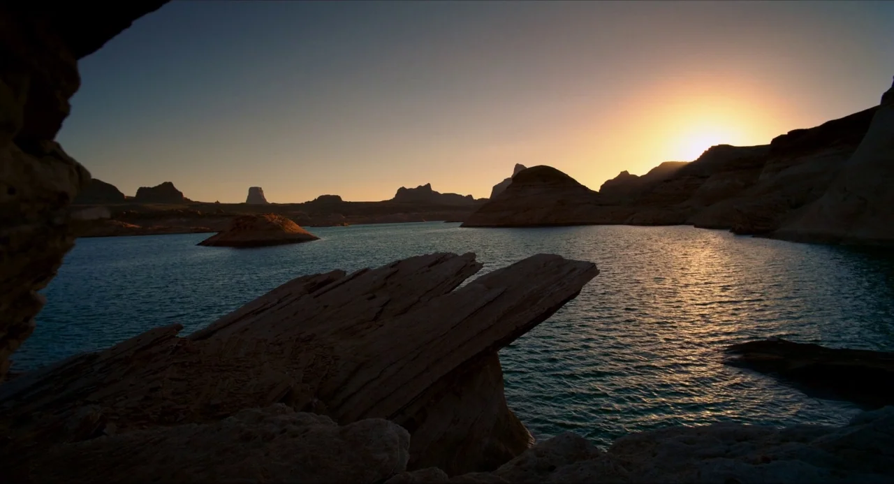 Voyage of Time (2016) movie still: establishing — A vast lake surrounded by desert rock formations and mesas at sunset. The golden sun rests just…
