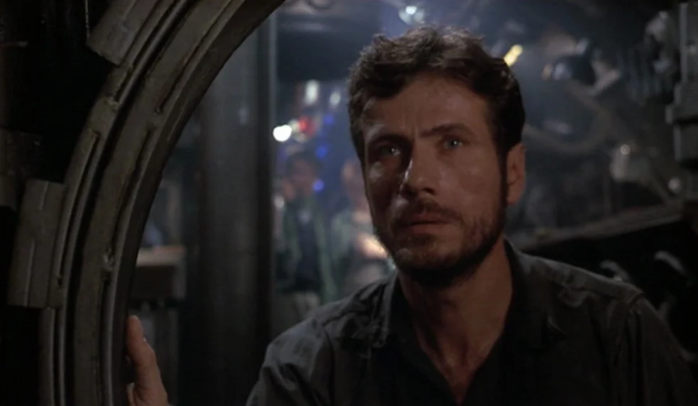 Das Boot (1981) movie still: medium — The interior of a cramped, dimly lit submarine with curved metal walls and pipes. A man with a beard leans…