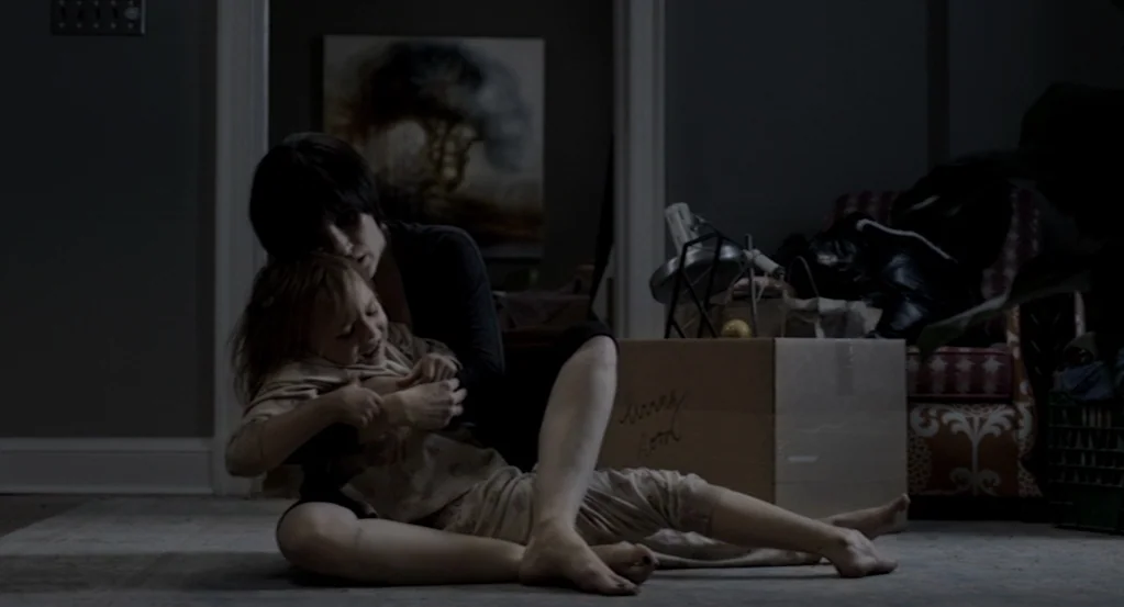 Mama (2013) movie still: medium — A dimly lit room contains cardboard moving boxes and a patterned armchair in shadows. A woman sits on the carpet…