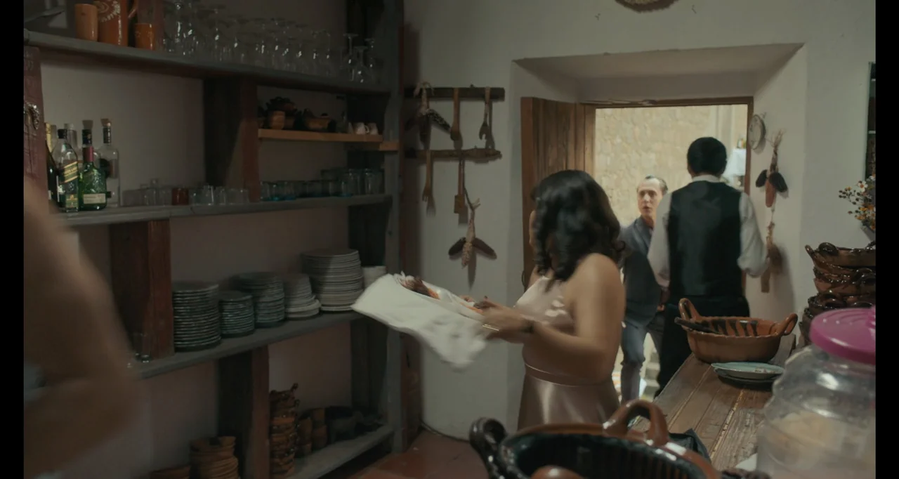 National Family (2023) movie still: medium — A restaurant back area contains wooden shelves stacked with plates and glassware. A woman in a satin…
