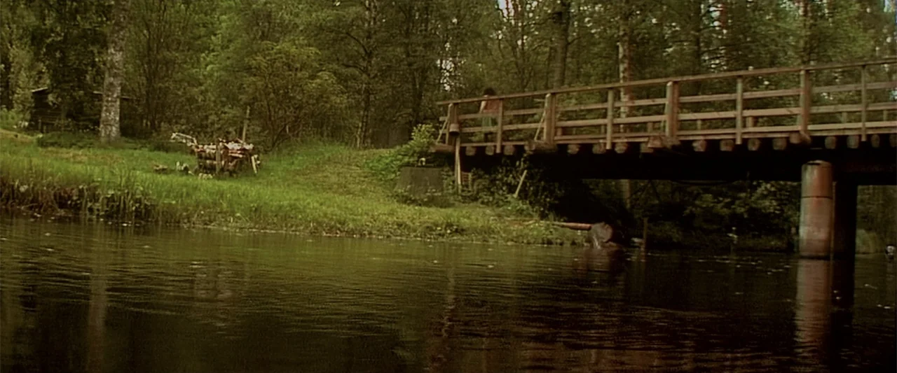 Dancer in the Dark (2000) movie still: wide — A lush forest area features a wooden bridge crossing over a still dark river. A woman stands on the…
