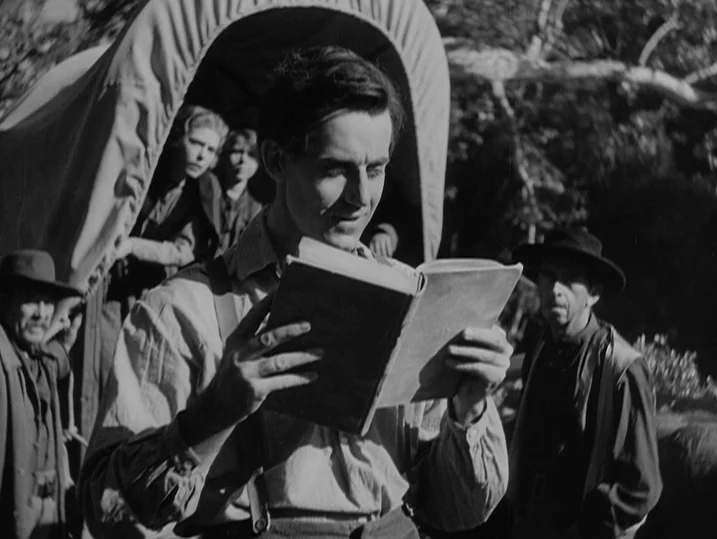 Young Mr. Lincoln (1939) movie still: medium — A group of people gathers near a covered wagon in a wooded setting. A young man reads from a book…