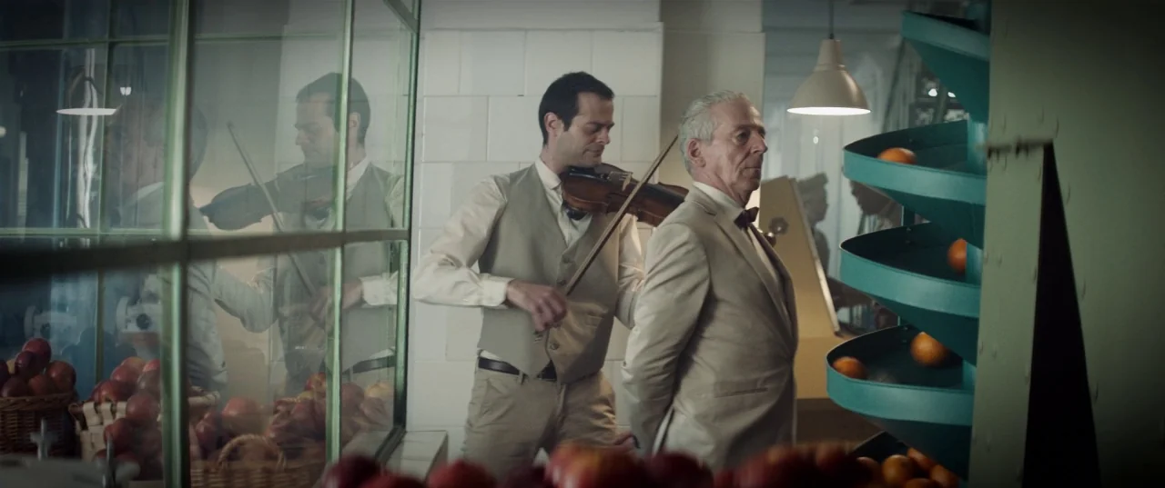 The Platform (2019) movie still: medium — A sterile industrial room contains a spiral metal sorting ramp and tiled walls. A man in a tan suit stands…