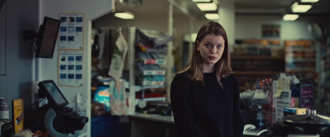 Rift (2017) movie still: medium — A interior view of a cluttered retail counter with information posters and digital terminals. A young woman stands…