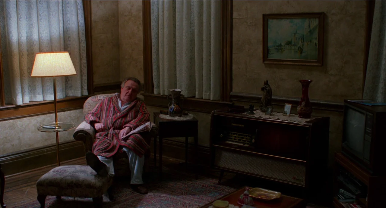 Moonstruck (1987) movie still: fullbody — An aging home interior with wood paneling and patterned wallpaper. A man sleeps in a chair while a lamp…
