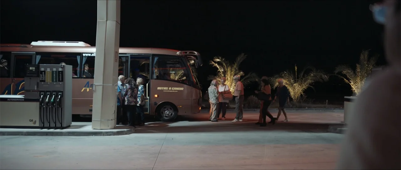 Historias lamentables (2021) movie still: wide — A night exterior at a gas station with a bus parked beneath canopy lights. A group of older adults…
