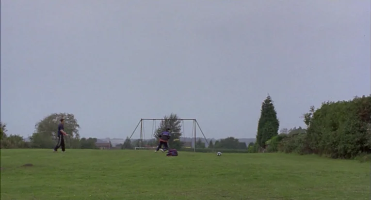 A Room For Romeo Brass (1999) movie still: wide — A green grassy playing field features a metal swing set under a grey sky. Two individuals move…