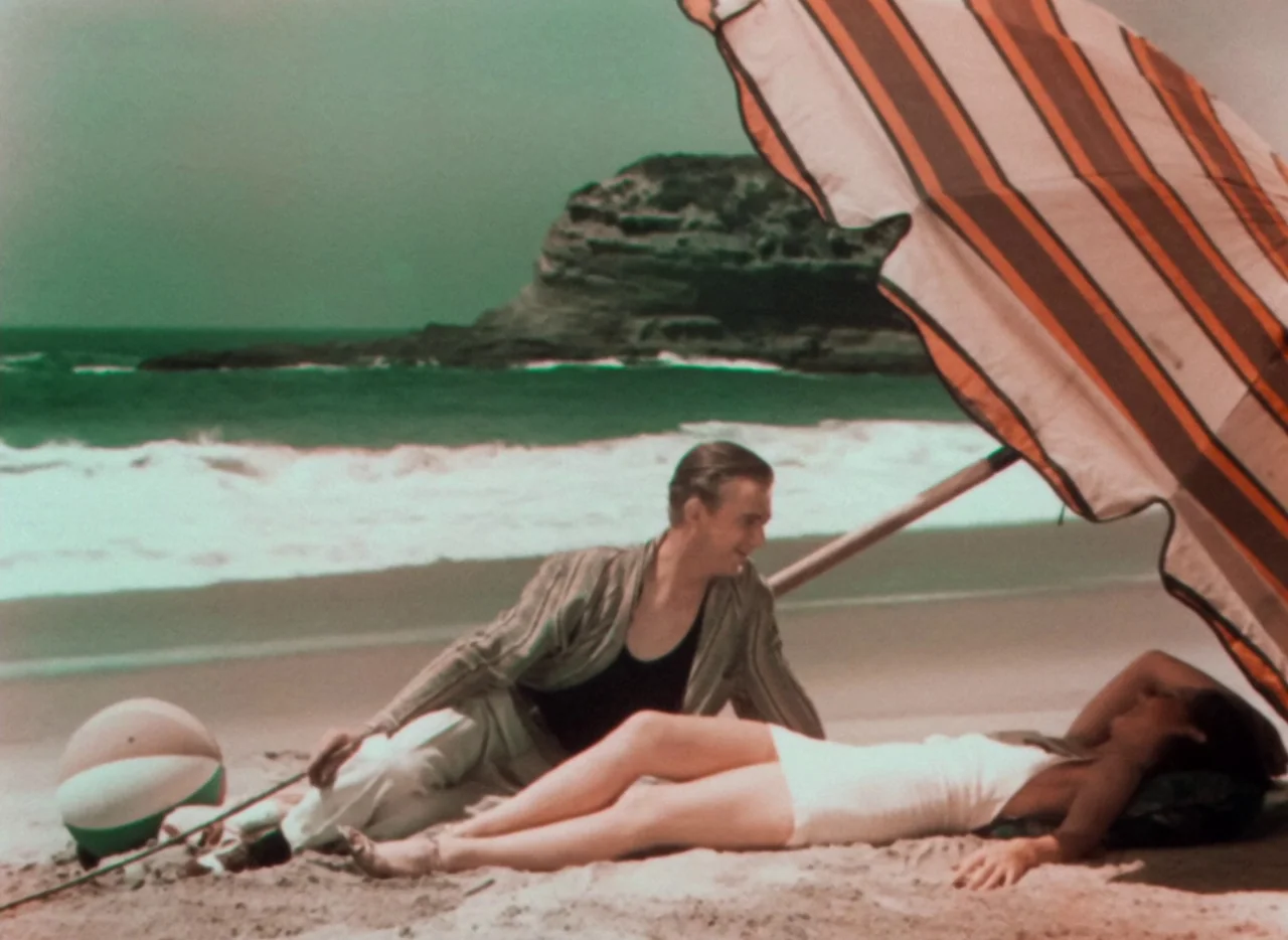 Doctor X (1932) movie still: medium — A sun-drenched beach featuring a sand dune, crashing surf, and an orange-striped umbrella. A man leans near a…