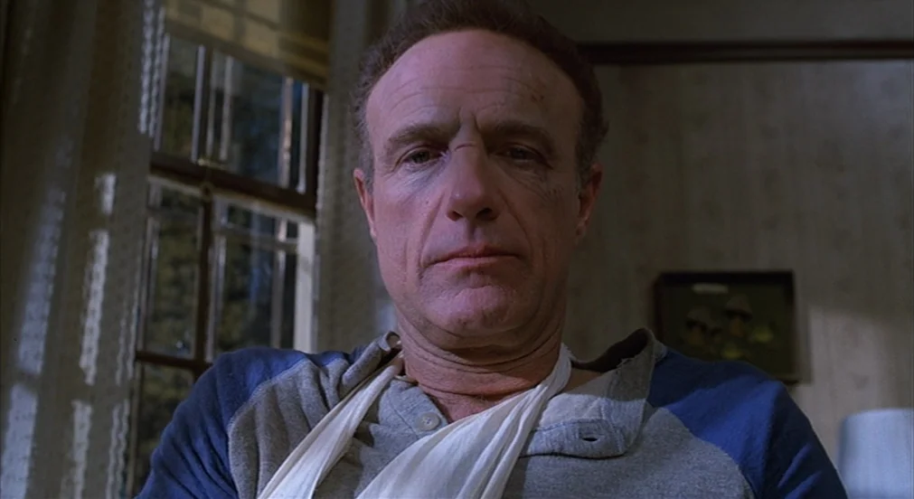 Misery (1990) movie still: closeup — A dimly lit room with wallpaper and a window with blinds in the background. A middle-aged man with a facial…