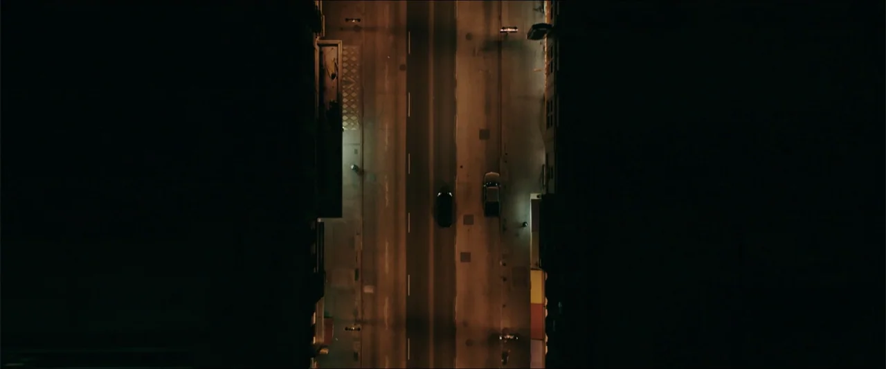Truth or Dare (2018) movie still: establishing — A vertical overhead perspective displays an urban canyon with two vehicles parked on asphalt. Deep…