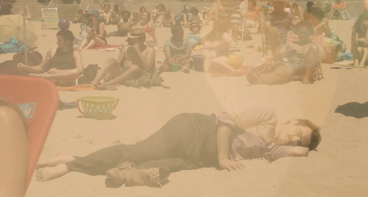 Her (2013) movie still: fullbody — A sunny public beach filled with people relaxing on the sand under bright daylight. A man in formal trousers and…