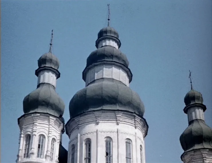 Viy (1967) movie still: establishing — Three white stone church towers with dark, onion-shaped domes rise toward a blue sky. The masonry displays…