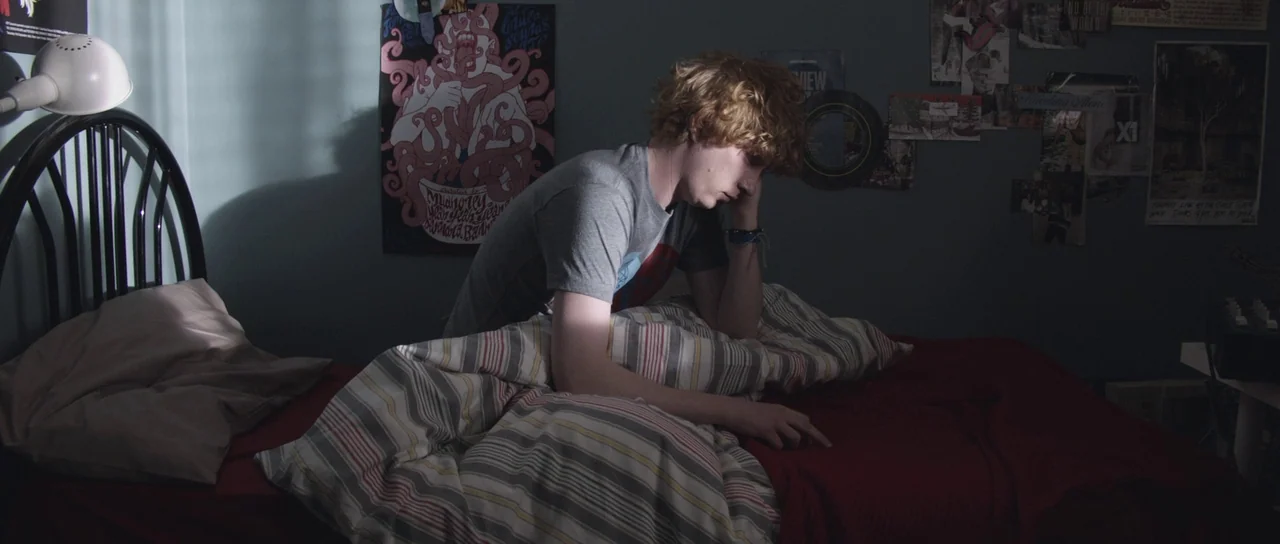 Frank (2014) movie still: medium — A dimly lit room with walls adorned with various music posters and collages. A young man with curly red hair sits…