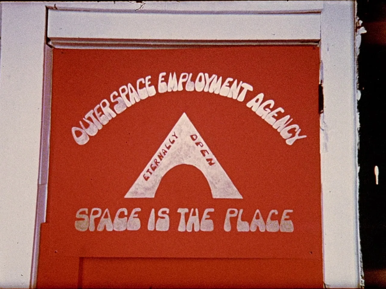 Space is the Place (1974) movie still: closeup — A flat red painted door panel featuring white hand-lettered text and a central arch. The sign reads…
