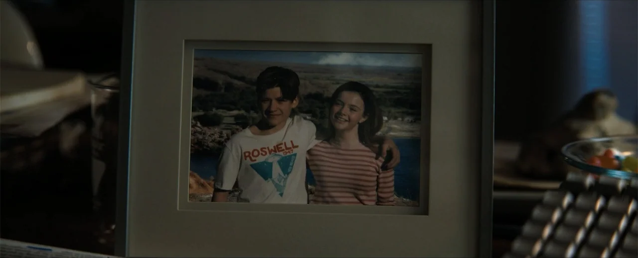 Venom: The Last Dance (2024) movie still: closeup — A framed photograph sits on a dark surface in a dimly lit room. The photo shows two teenagers…