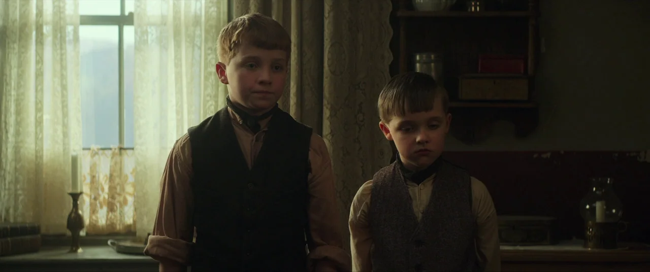 Sunset Song (2015) movie still: medium — Two young boys stand before a lace-curtained window in an austere, sparsely furnished room. They wear…