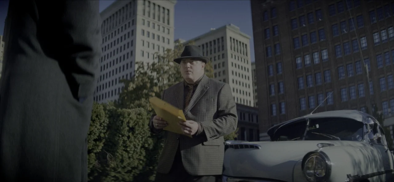 No Sudden Move (2021) movie still: medium — A man stands on a city street next to a vintage light-colored car. He holds a yellow envelope while…