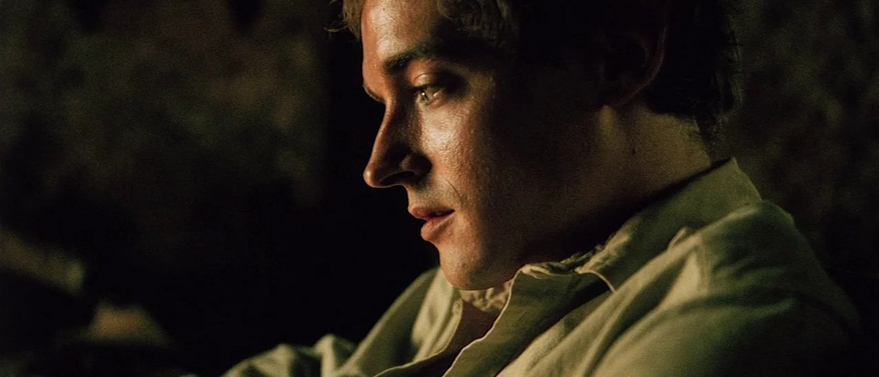 Renegade (2004) movie still: closeup — A man in a cream-colored shirt sits in a dimly lit, dark-toned interior space. He looks downward while his…