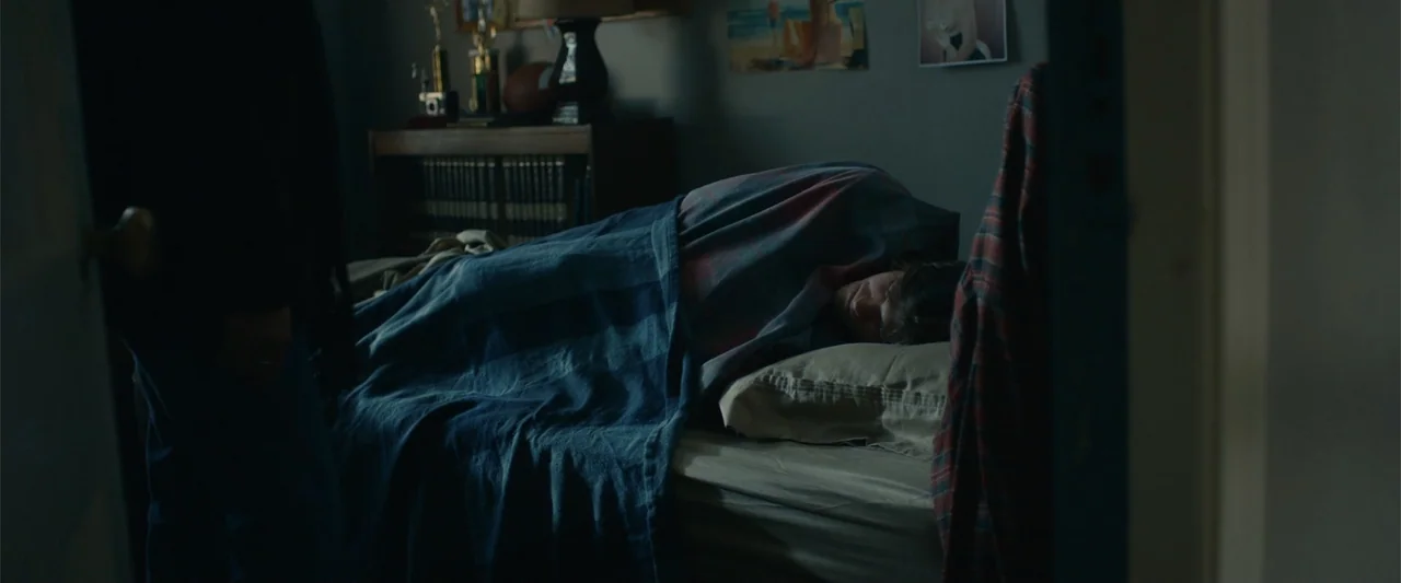 Coming Through the Rye (2016) movie still: medium — A dimly lit room contains a bed with a person sleeping under a blanket. A figure stands in the…
